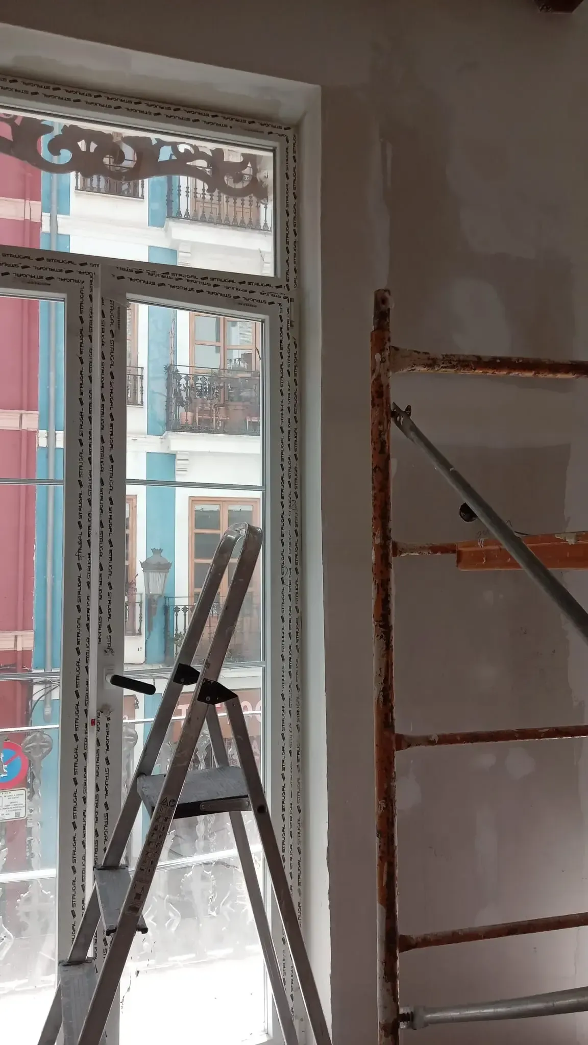 Habitación interior en obras, con andamios y una ventana abierta con vistas a los edificios del exterior.