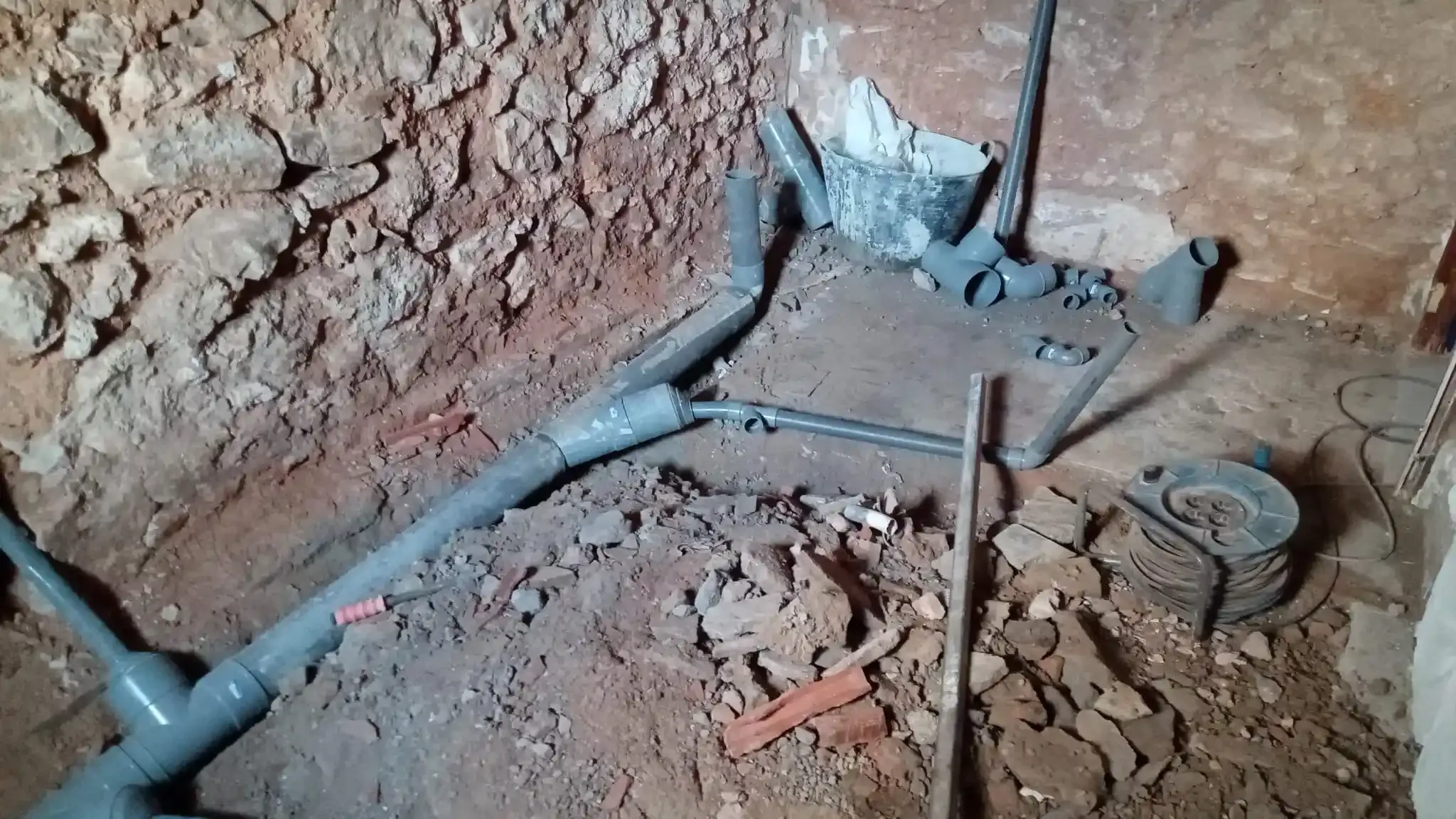 Tuberías grises rotas en una zanja de tierra junto a un muro de tierra, con juntas de fontanería expuestas y escombros.