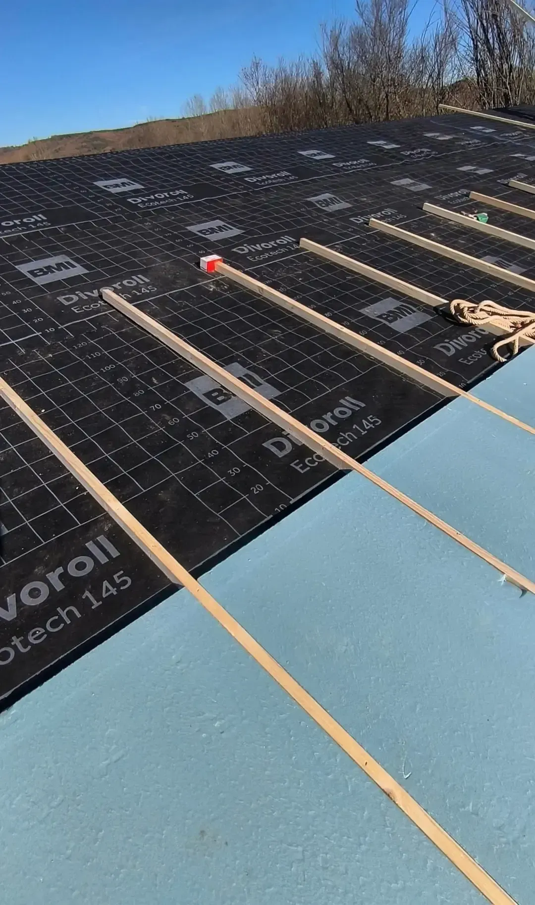 Hombre con camisa y gorra azules, reparando tejas de barro en un tejado, utilizando herramientas.
