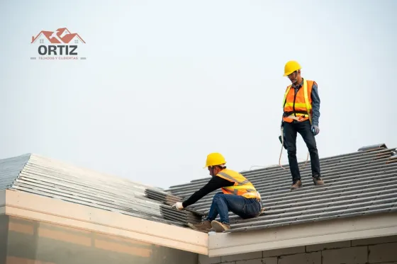 Dos techadores con cascos y chalecos antibalas instalando tejas en una casa bajo un cielo despejado.