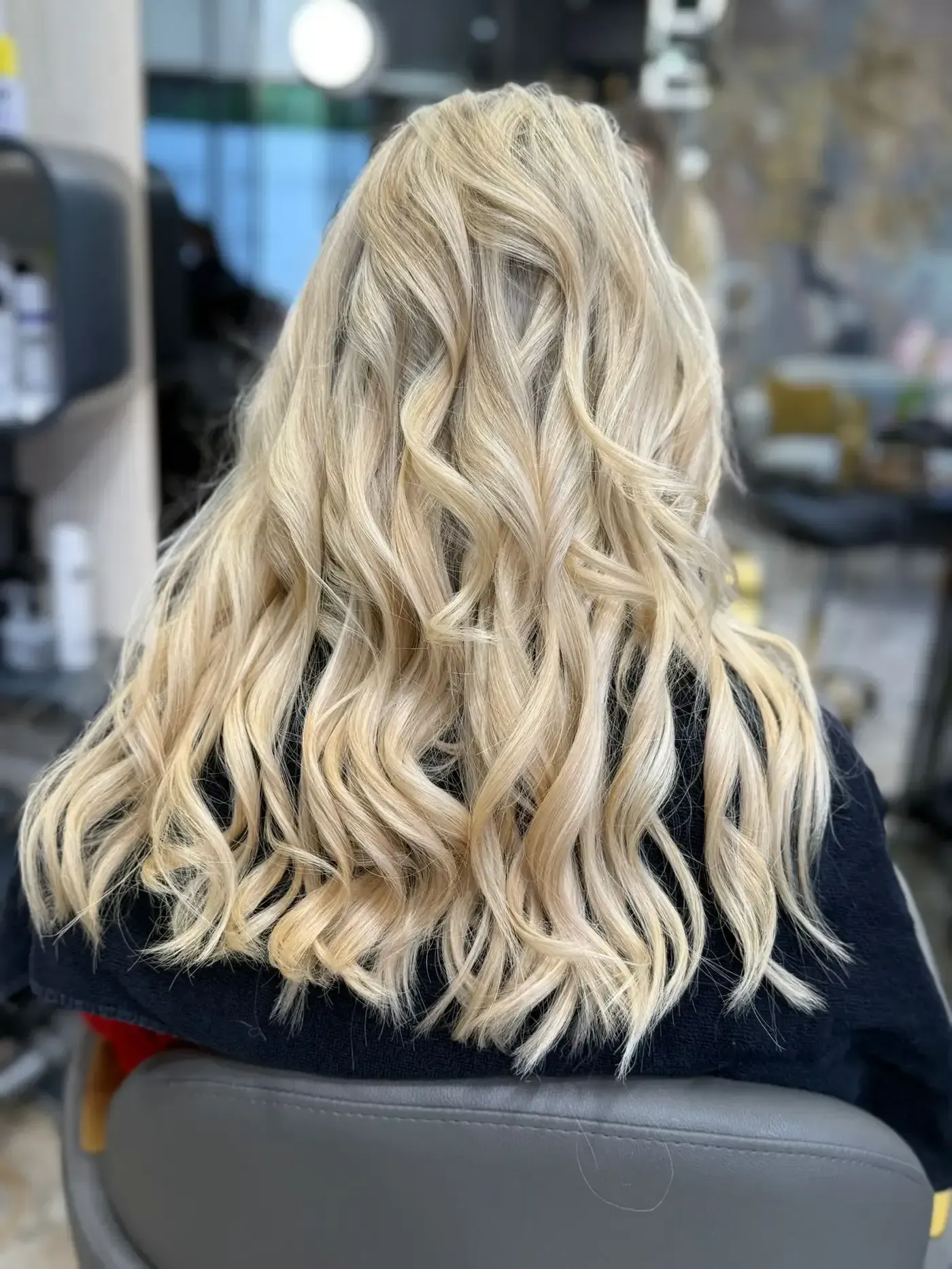 Cabello rubio claro y ondulado peinado sobre los hombros de una persona, sentada en una silla de salón.