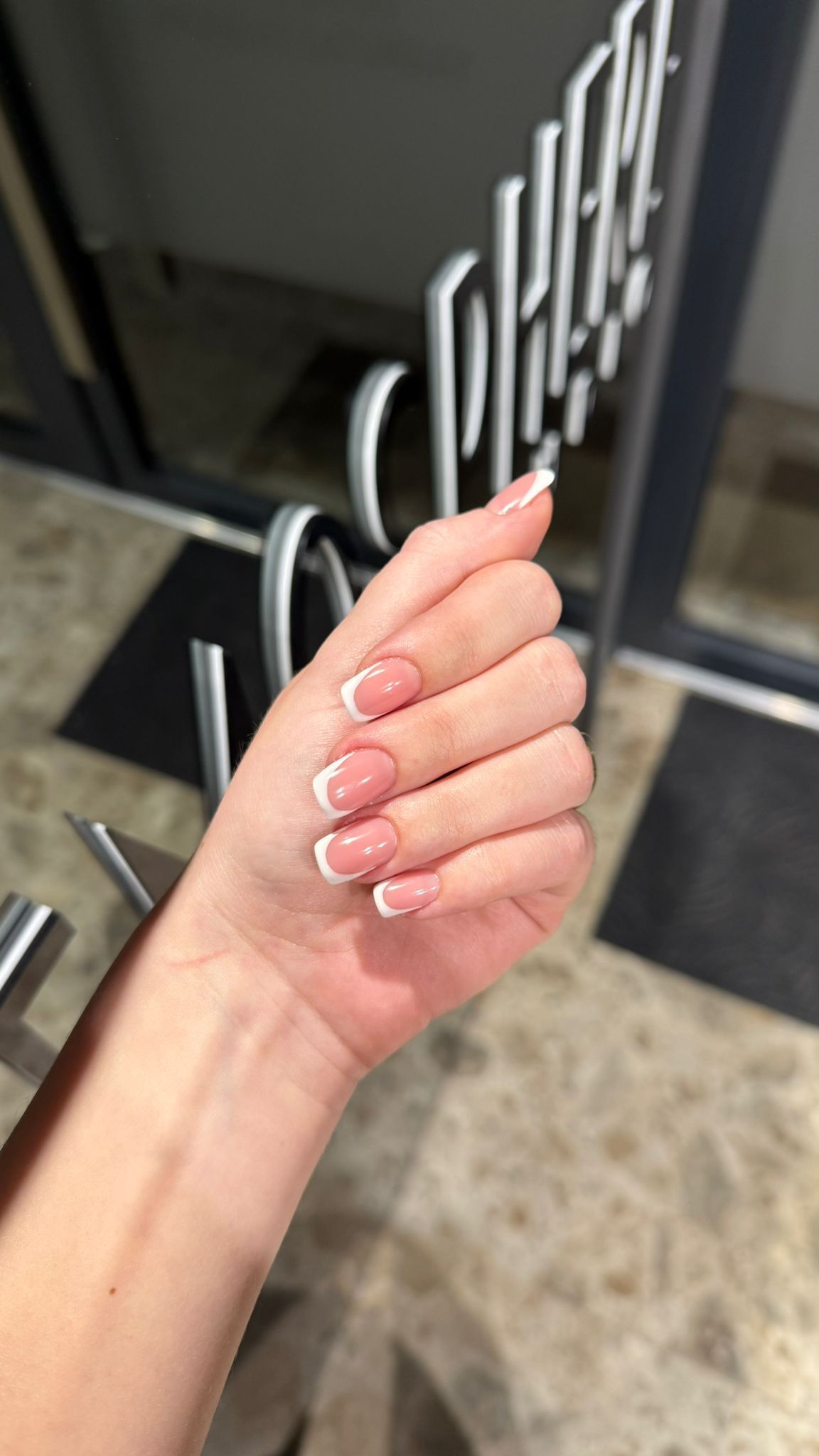 Mano con uñas de punta francesa, posando en un interior cerca de una puerta y un tapete.