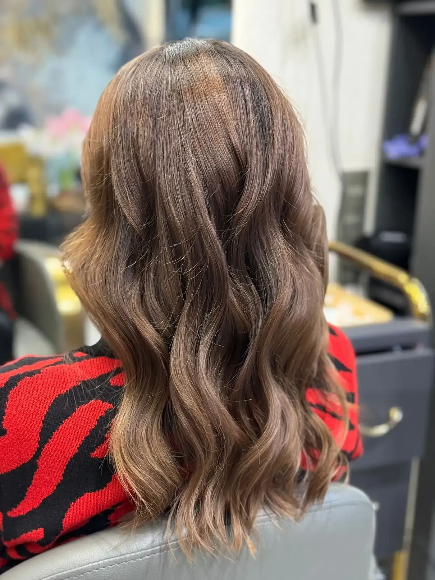 Mujer con cabello largo castaño y ondulado, vistiendo un suéter rojo y negro, en una silla de salón.