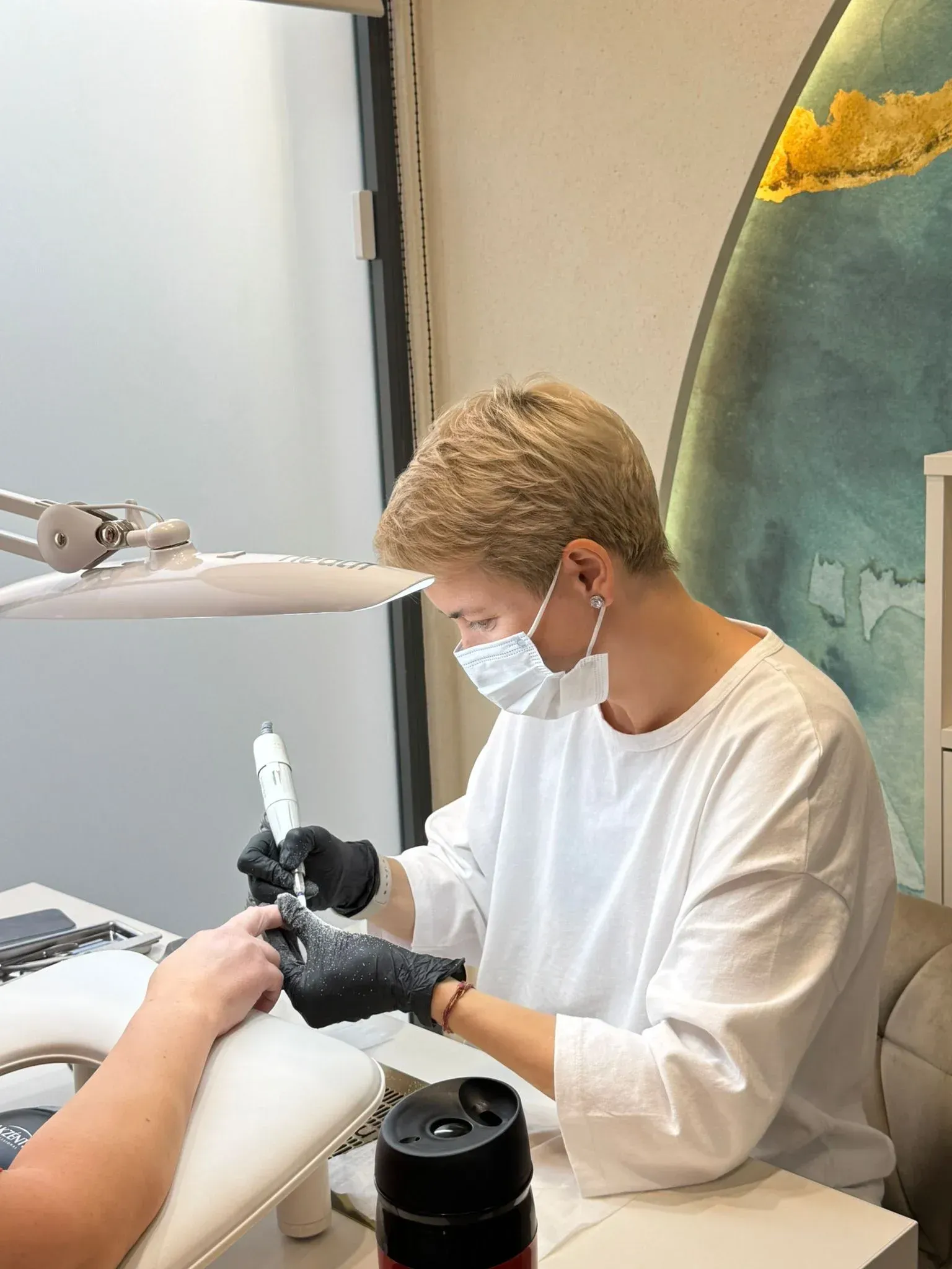 Persona con mascarilla aplicándose esmalte de uñas en un salón. Lleva guantes negros y camisa blanca.