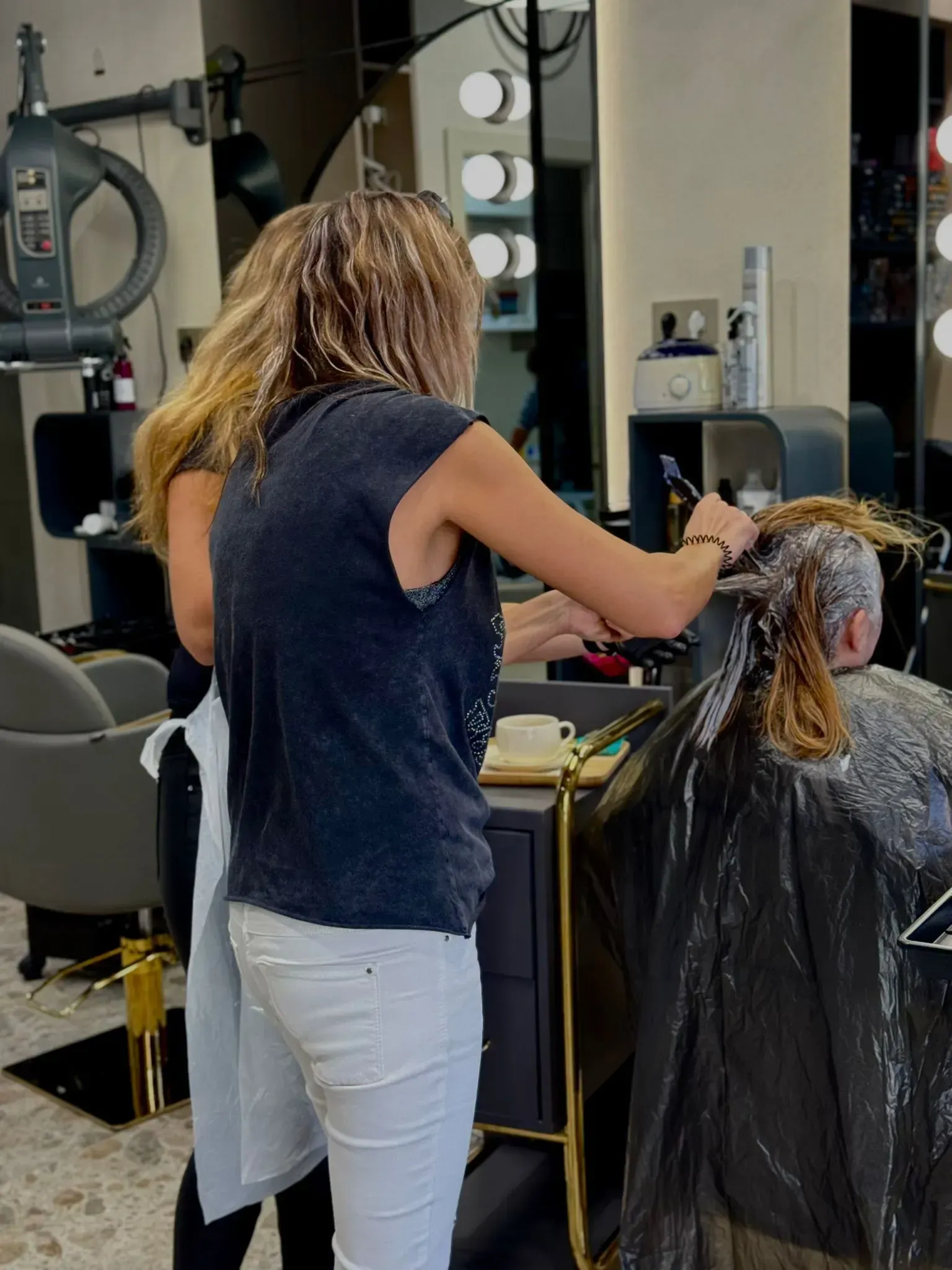 Peluquera aplicando tinte a una clienta en un salón. Capa negra, pantalón blanco, espejo y luces visibles.