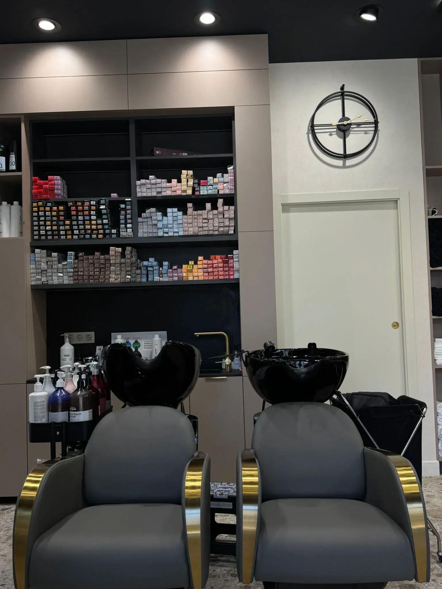 Peluquería con dos sillas para champú, productos en estantes y reloj en la pared.