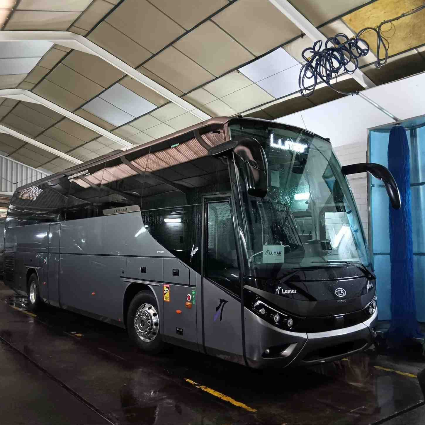 Un autobús turístico Lumar de color gris oscuro estaba estacionado en un terreno de grava frente a una iglesia.
