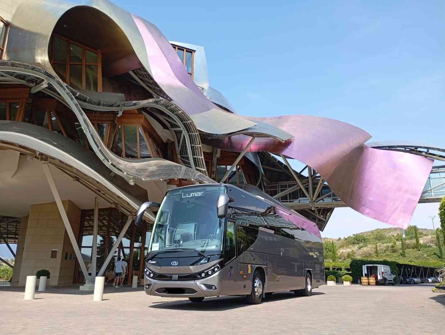Un elegante autobús turístico de color gris oscuro estaba estacionado frente a la arquitectura curvilínea.