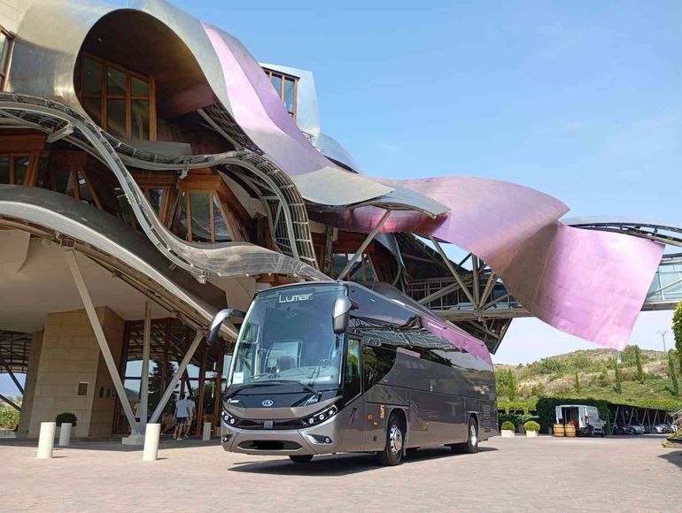 Un elegante autobús turístico de color gris oscuro estaba estacionado frente a la arquitectura curvilínea.