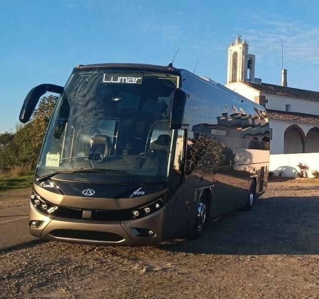 Un autobús turístico Lumar de color gris oscuro estaba estacionado en un terreno de grava frente a una iglesia.