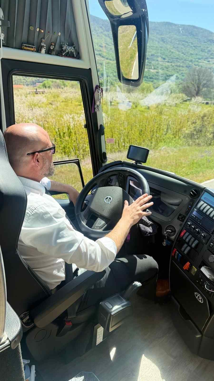Una persona con camisa azul claro abotonada y corbata azul conduciendo un autobús, con las manos en el volante.