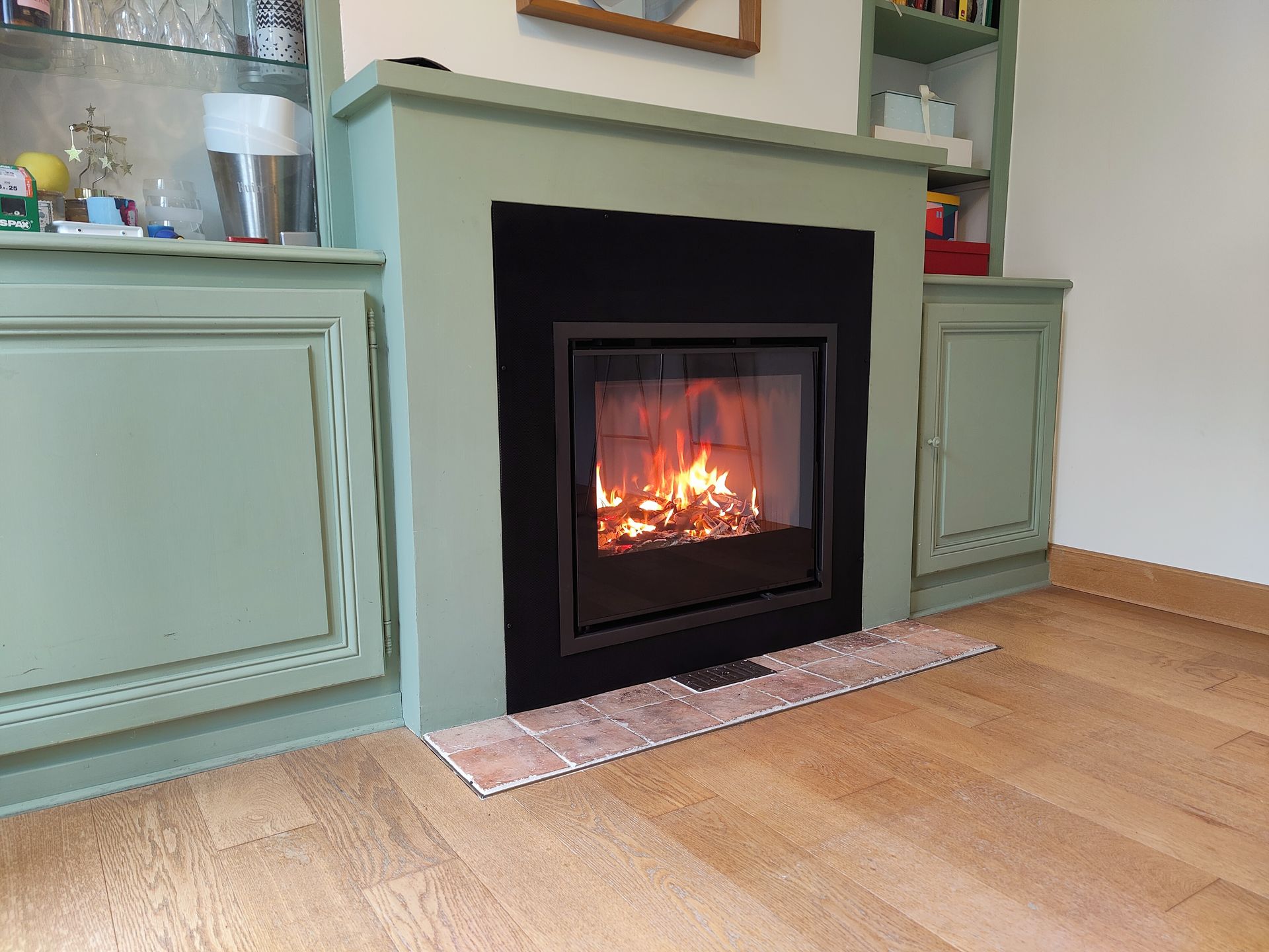 Cheminée encastrée verte avec un feu crépitant dans une pièce claire au parquet en bois