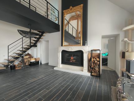 Salon moderne avec escalier noir, cheminée blanche, grand miroir, parquet et plafond à poutres apparentes