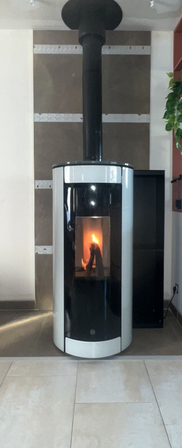 Poêle à bois moderne avec cheminée noire dans une pièce carrelée, le feu est visible à l'intérieur.