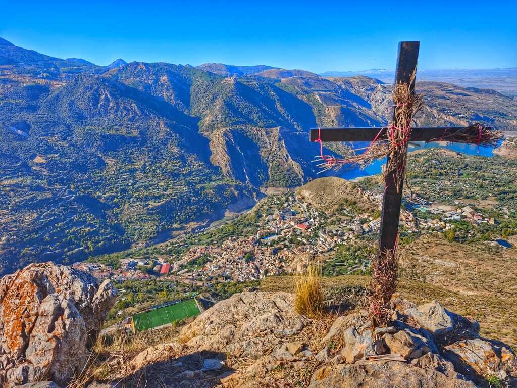 Una cruz en la cima de una colina rocosa con vistas a la montaña, un lago y un pueblo a sus pies.