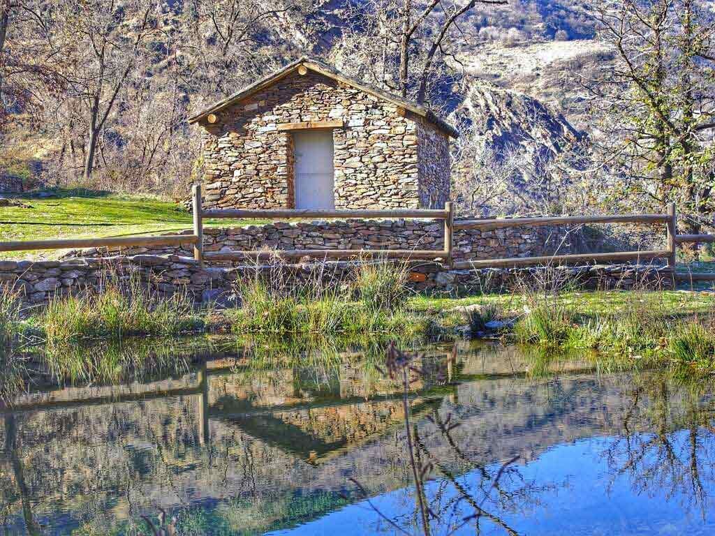 Cabaña construida con leña junto a un estanque reflectante; montañas al fondo.