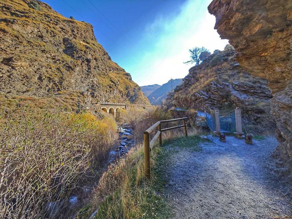 Vista panorámica de un cañón con un sendero, un puente y coches a lo largo de un río bajo un cielo azul.