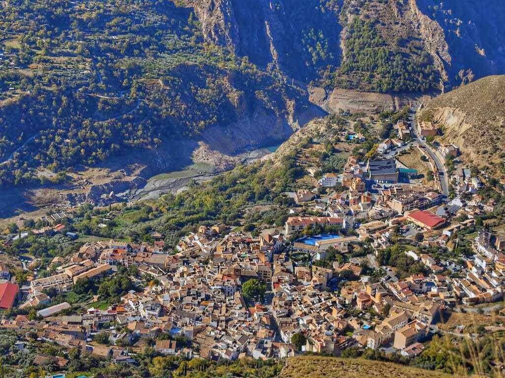 Vista aérea de un pueblo enclavado en un valle, rodeado de montañas y árboles. Día soleado, con edificios de colores variados.
