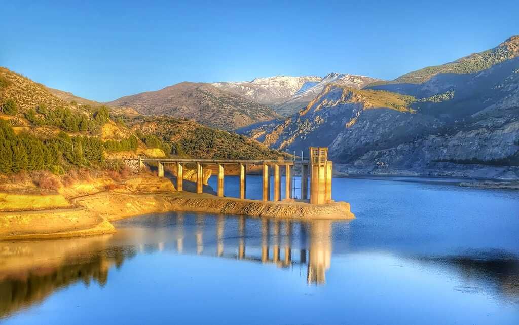 Paisaje lacustre con ruinas de un antiguo puente, montañas y agua azul que refleja el cielo.
