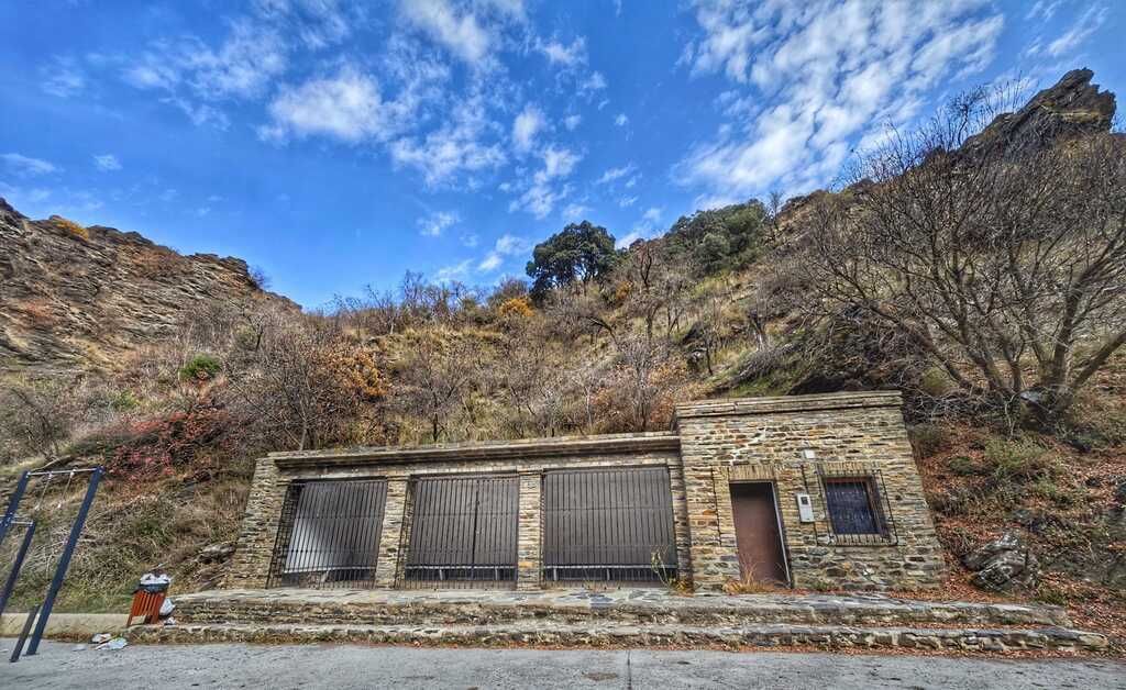 Edificio de piedra con una zona cubierta, junto a una colina rocosa, con un cielo azul de fondo.