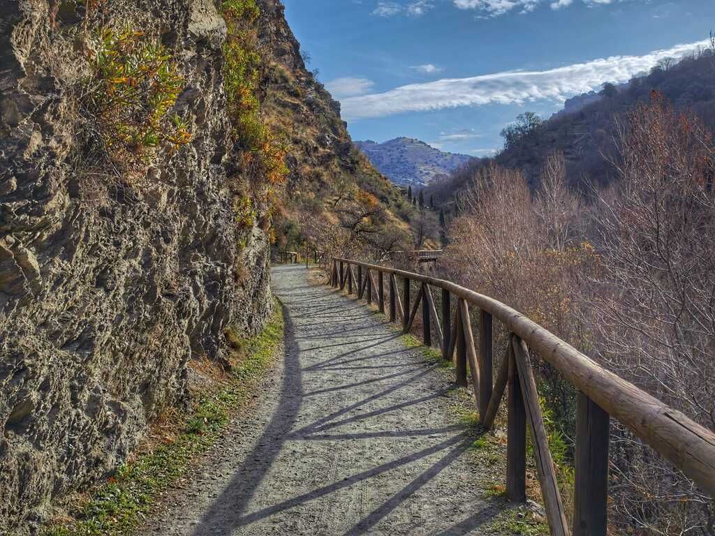 Un sendero serpentea a través de un desfiladero de montaña, con una valla de madera y acantilados rocosos a ambos lados bajo un cielo azul.