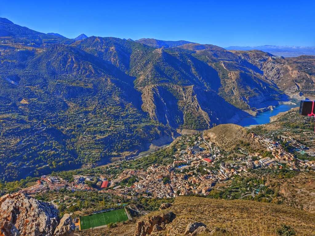 Paisaje de montaña con un pueblo a sus pies, un lago y un cielo azul despejado.