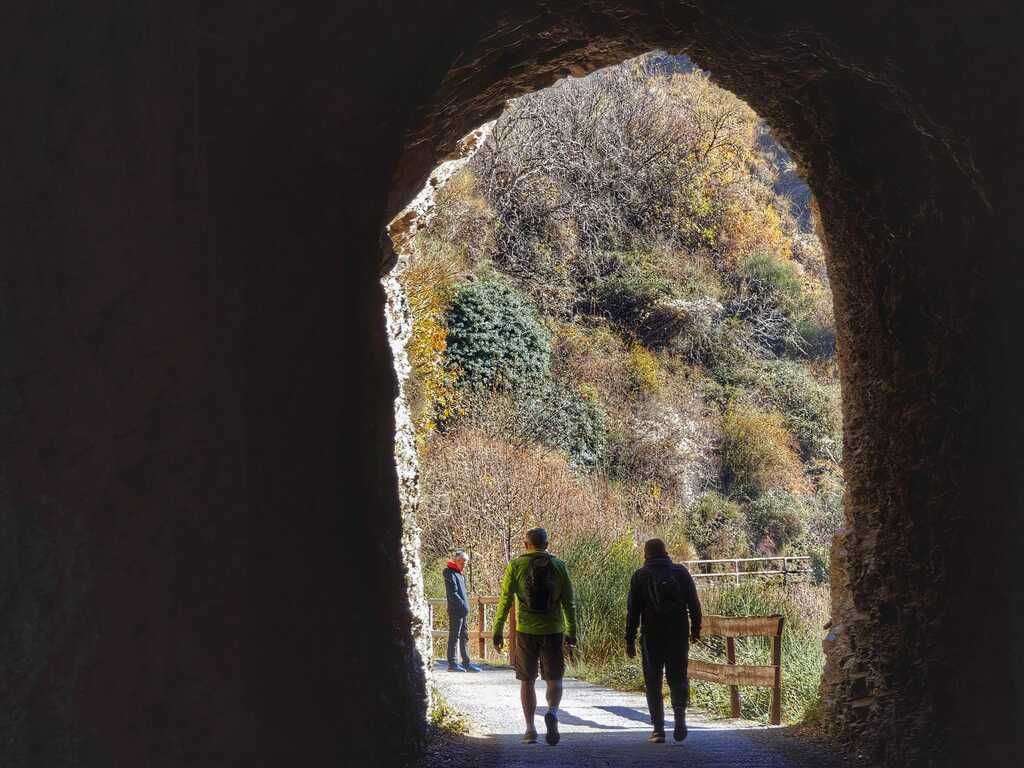 Personas caminando hacia un paisaje iluminado a través de la abertura de un túnel oscuro; árboles con colores otoñales.