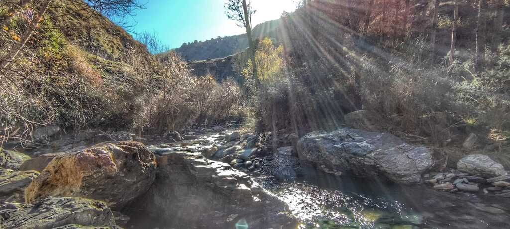 La luz del sol se filtra a través de un paisaje montañoso de rocas, un arroyo y matorrales secos en una ladera.