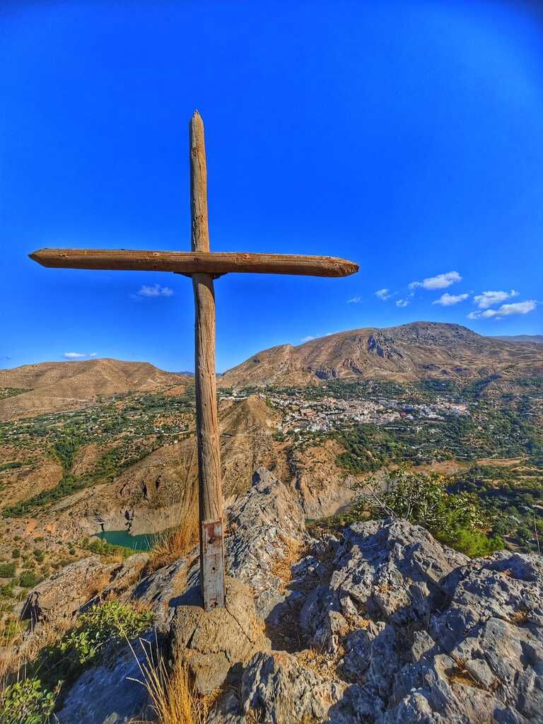Cruz de madera en lo alto de un pico rocoso con vistas a un pueblo, con montañas y cielo azul al fondo.