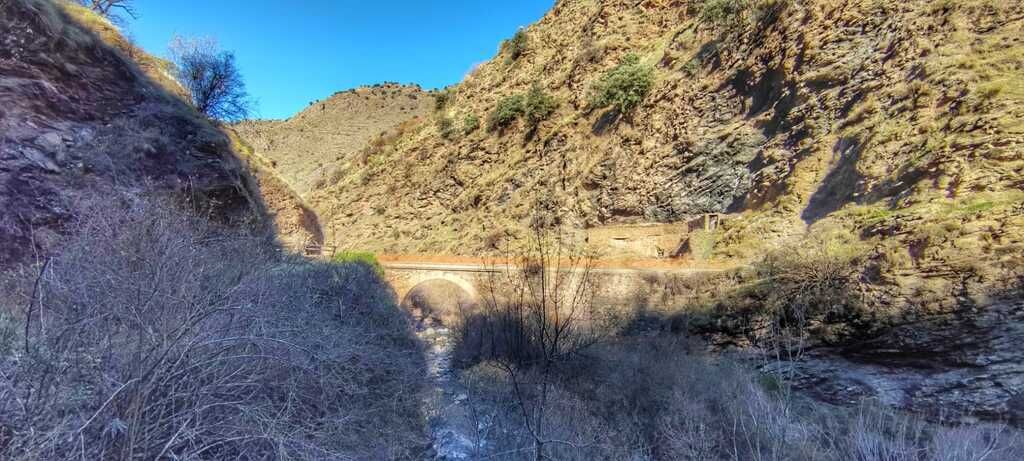 Paisaje montañoso con un puente que cruza un cañón; laderas marrones, cielo azul.