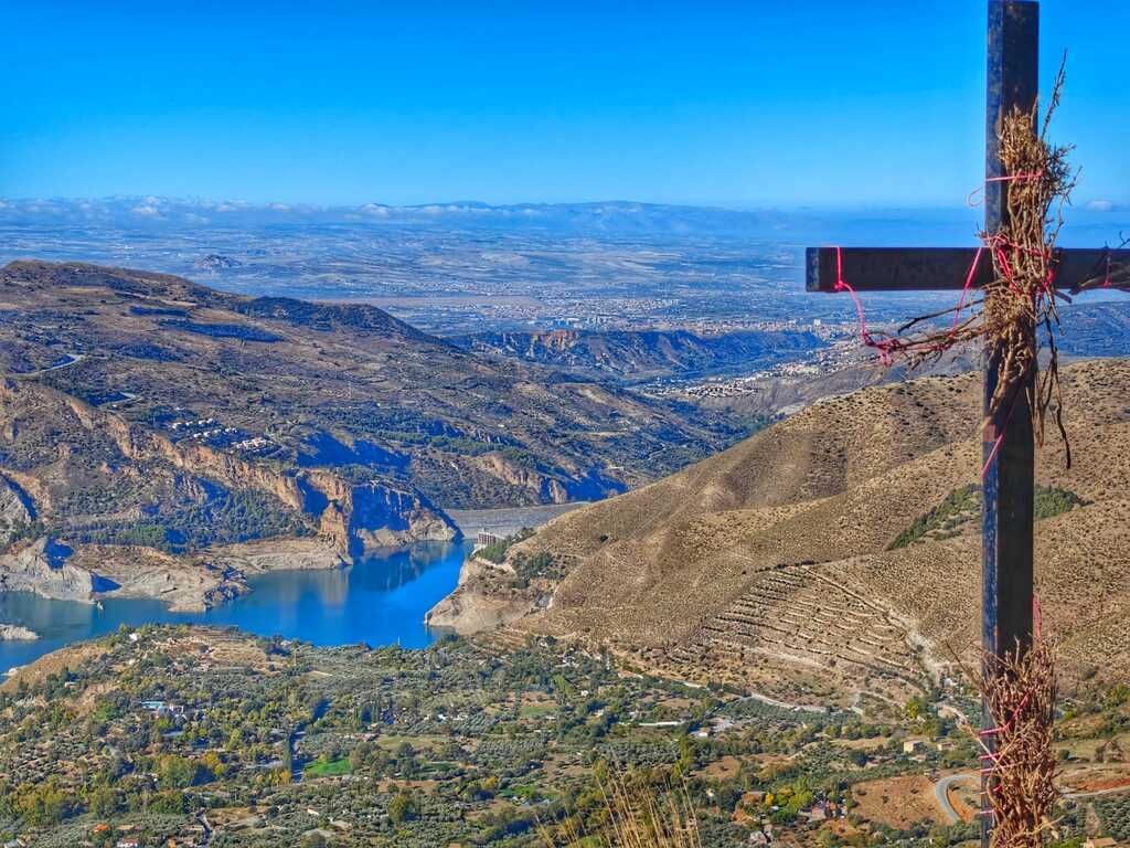 Vista desde la cima de una montaña con una cruz y un vasto paisaje con un lago y una ciudad a lo lejos.