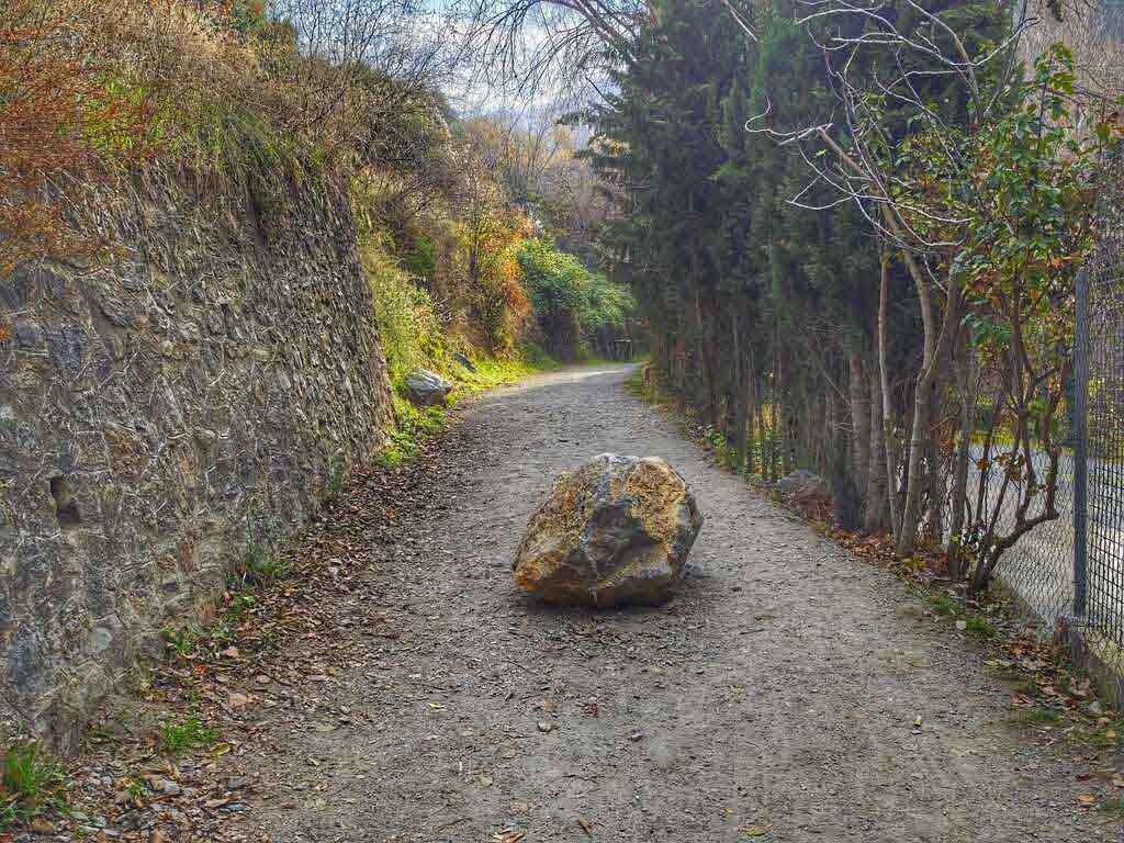 Una gran roca bloquea un camino de grava que serpentea por una ladera bordeada de árboles y un muro de piedra.