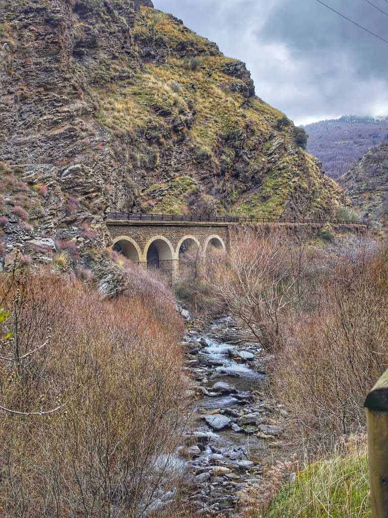 Puente de piedra con arco sobre un arroyo en un desfiladero de montaña. Vegetación árida y cielo nublado.