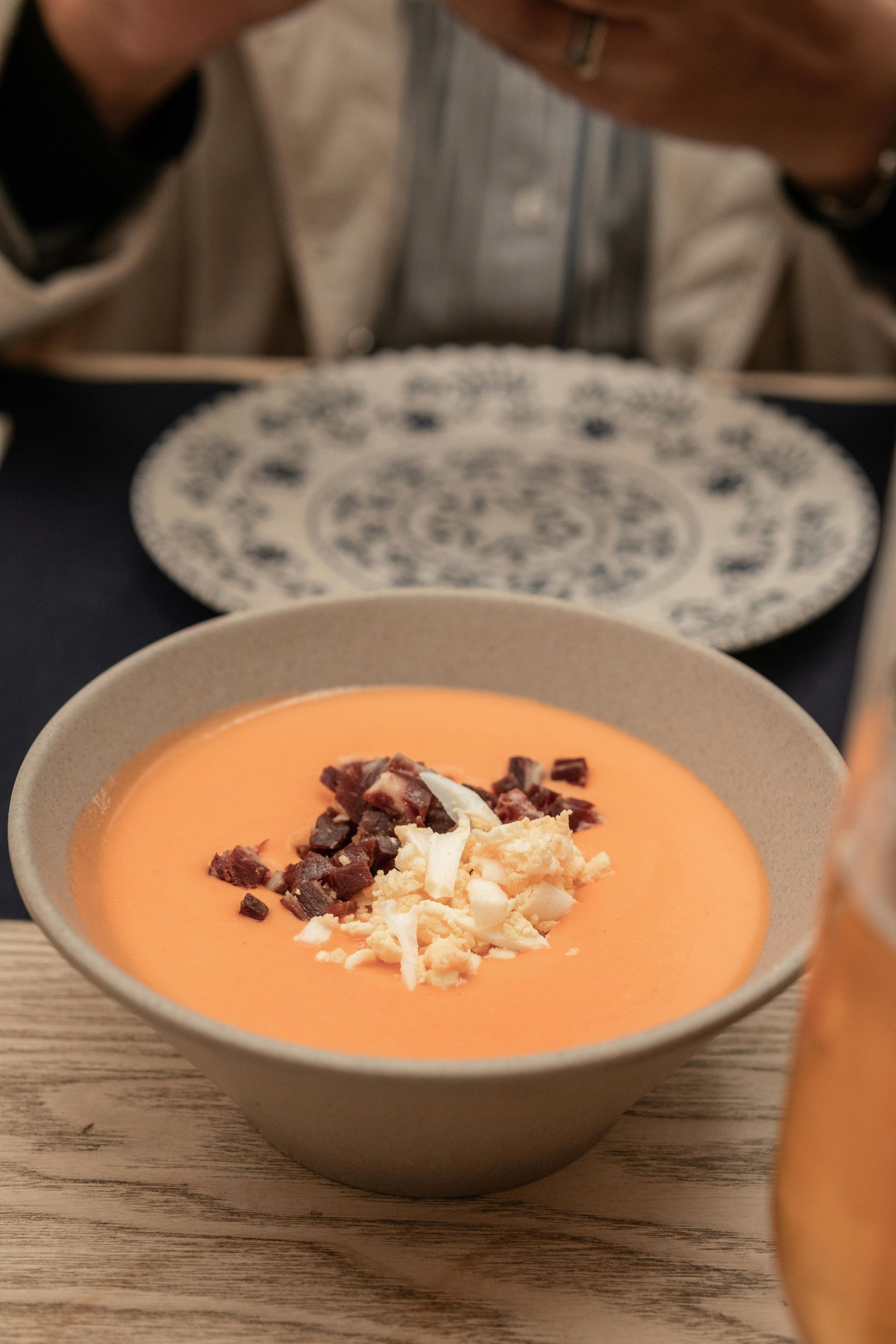 Sobre una mesa hay un tazón de sopa de gazpacho de naranja con jamón y huevo.