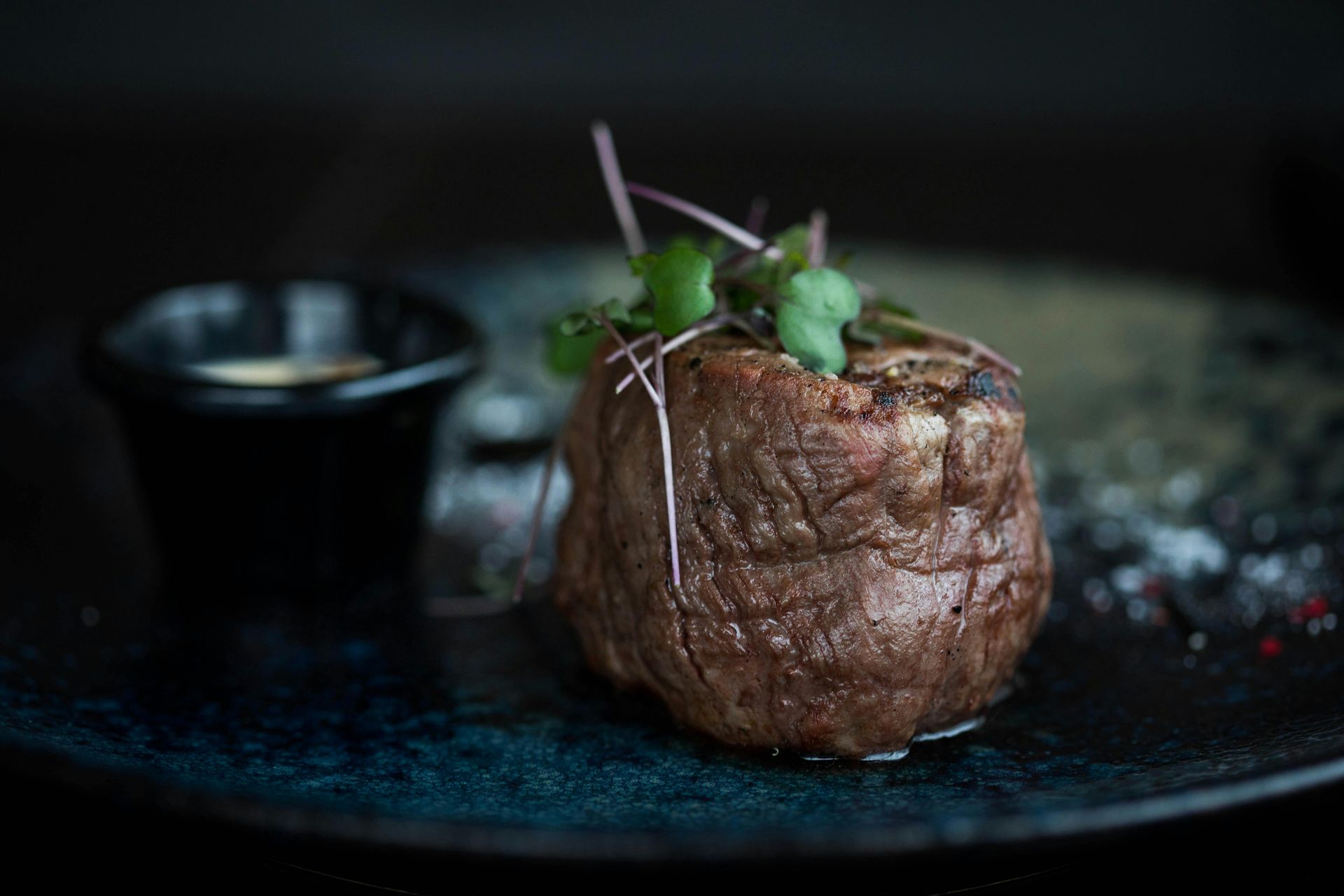 Bistec sellado con guarnición y un tazón pequeño de salsa en un plato azul oscuro.