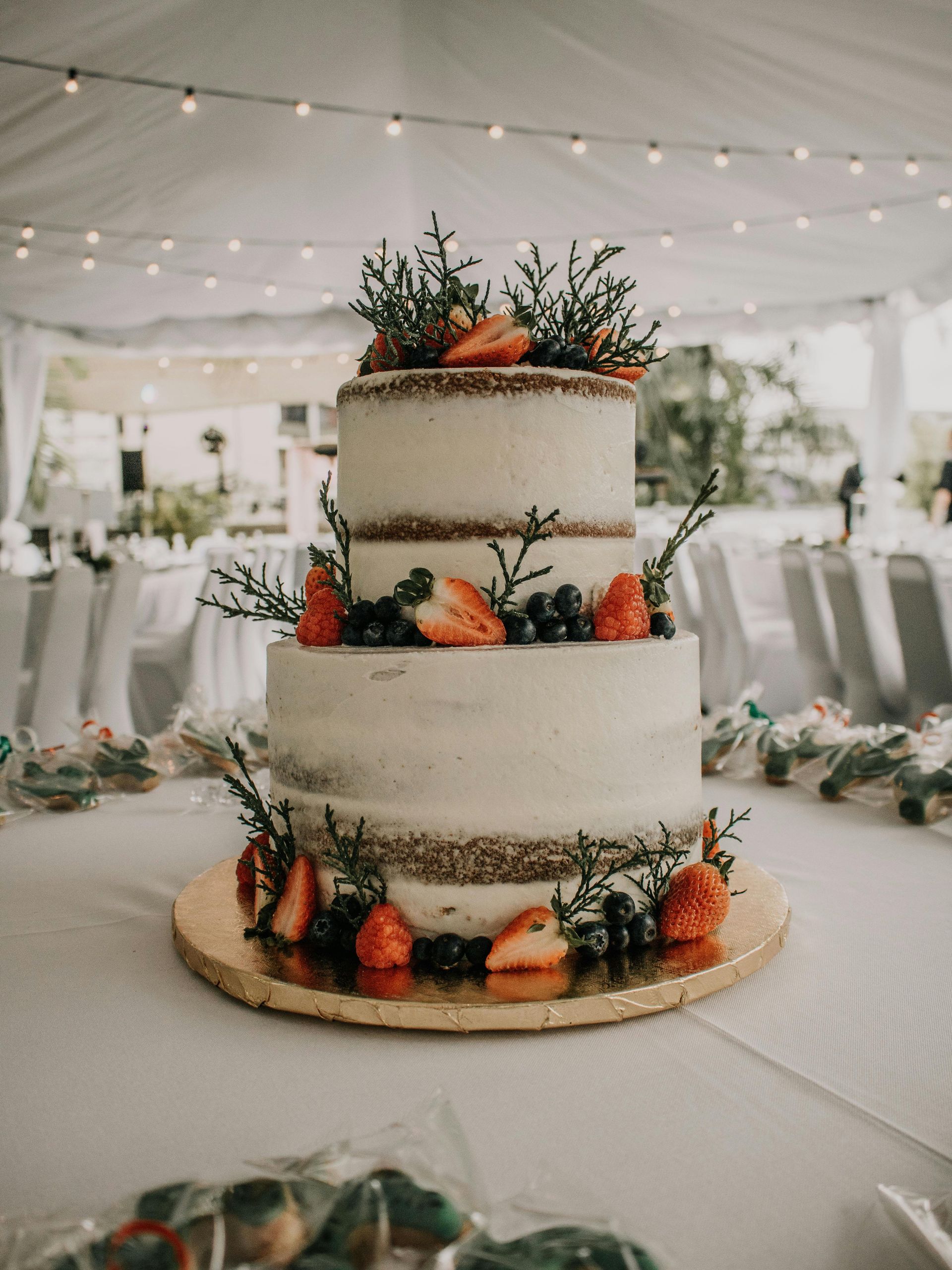 Pastel rústico de tres niveles decorado con fresas, arándanos y vegetación, sobre un soporte dorado