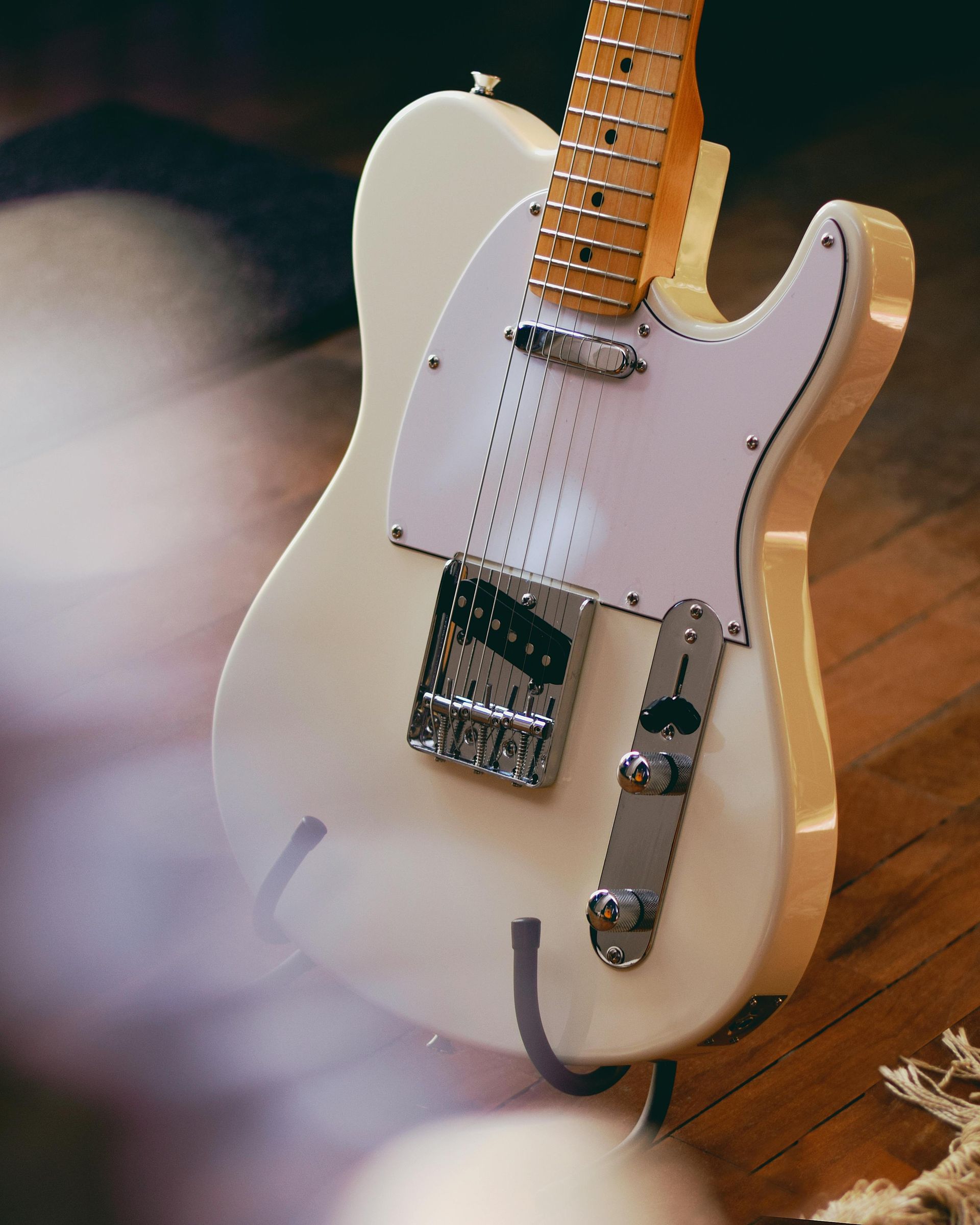 Guitarra eléctrica de color crema con golpeador blanco sobre soporte, fondo de piso de madera.