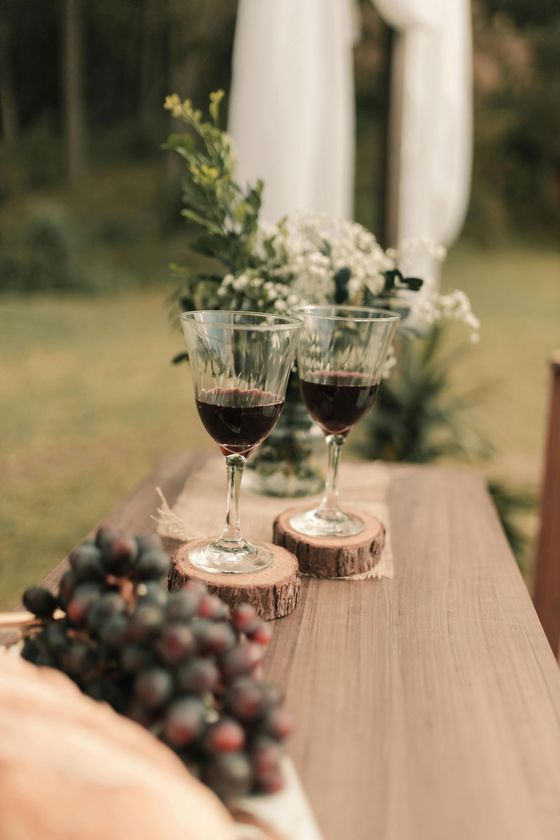 Dos copas de vino con vino tinto descansan sobre posavasos de madera junto a un racimo de uvas