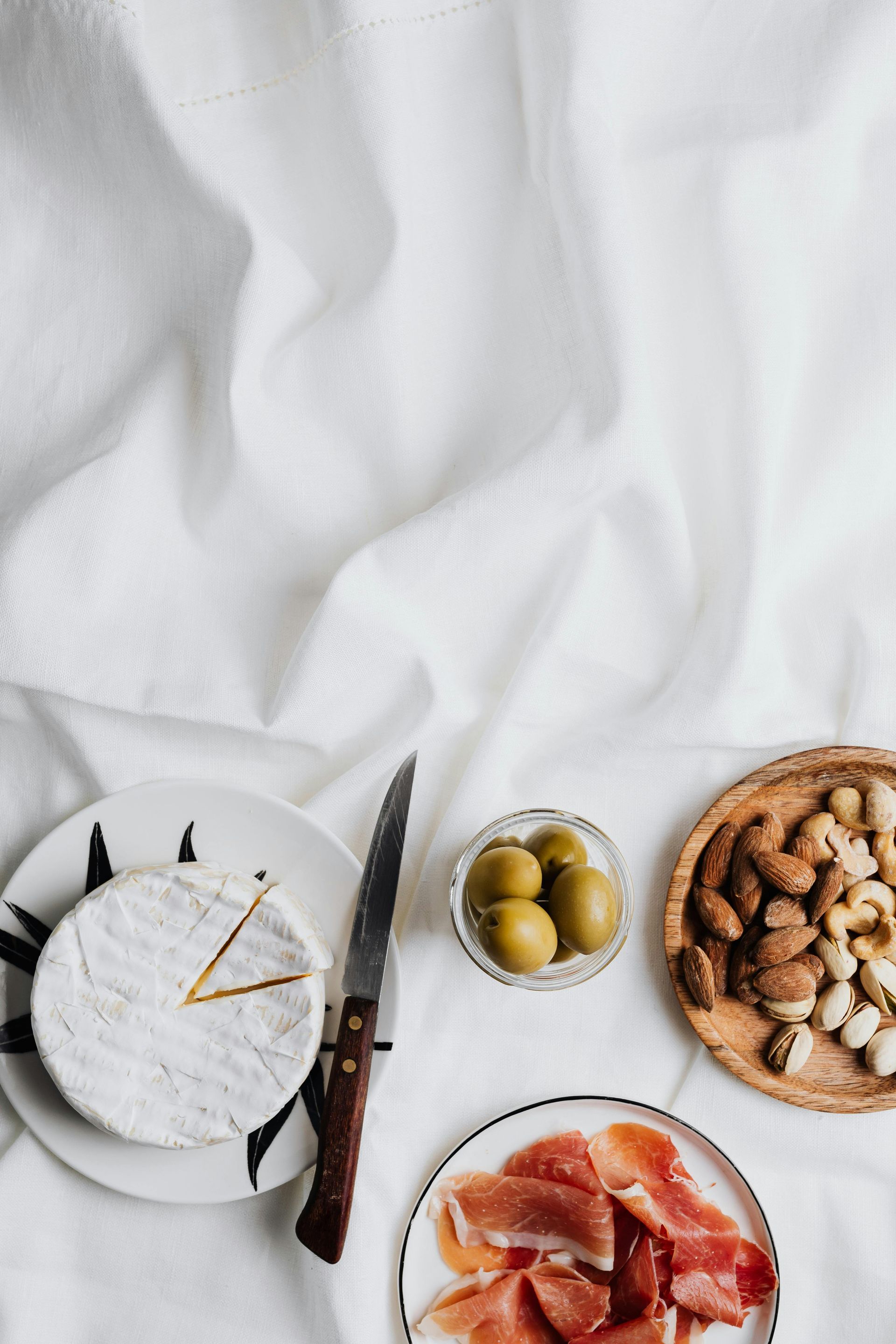 Queso, aceitunas, nueces y prosciutto sobre un paño blanco, con un cuchillo.