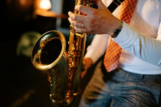 Hombre tocando un saxofón dorado, vistiendo una camisa blanca y corbata estampada, en un entorno poco iluminado.