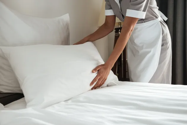Una persona con uniforme de hotel coloca una almohada blanca sobre una cama impecablemente hecha.