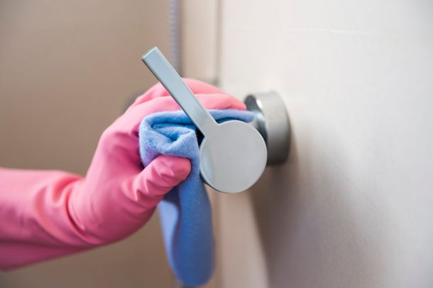 Una mano enguantada con un guante de goma rosa limpia el pomo metálico de una puerta con un paño de microfibra azul.
