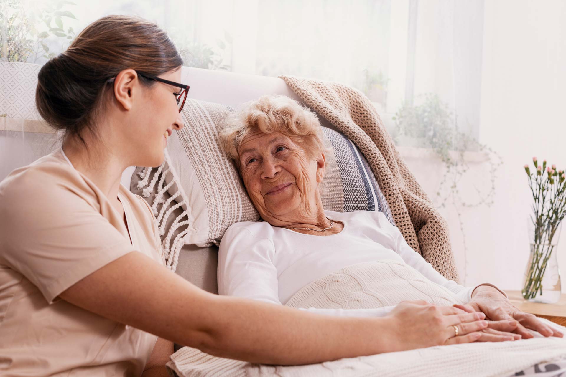Une personne âgée souriante assise dans son fauteuil, une aide soignante à son chevet lui souriant en lui prenant la main