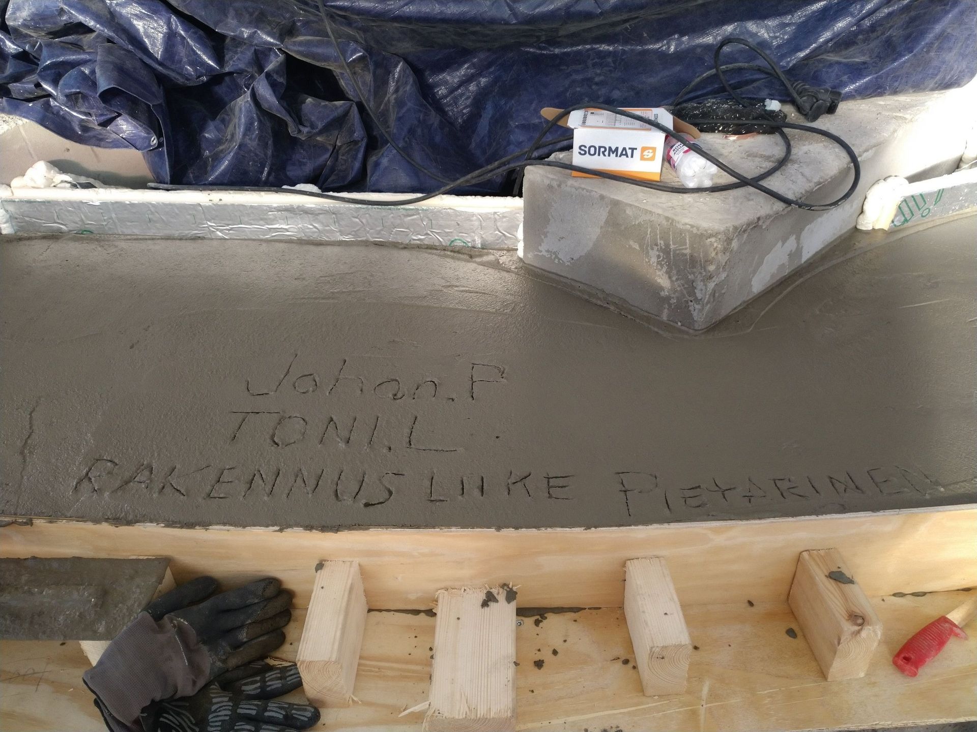 Tuore betonilaatta, jossa on haaleaa tekstiä, puumuotit, käsineet ja muurauslasta rakennustyömaalla.