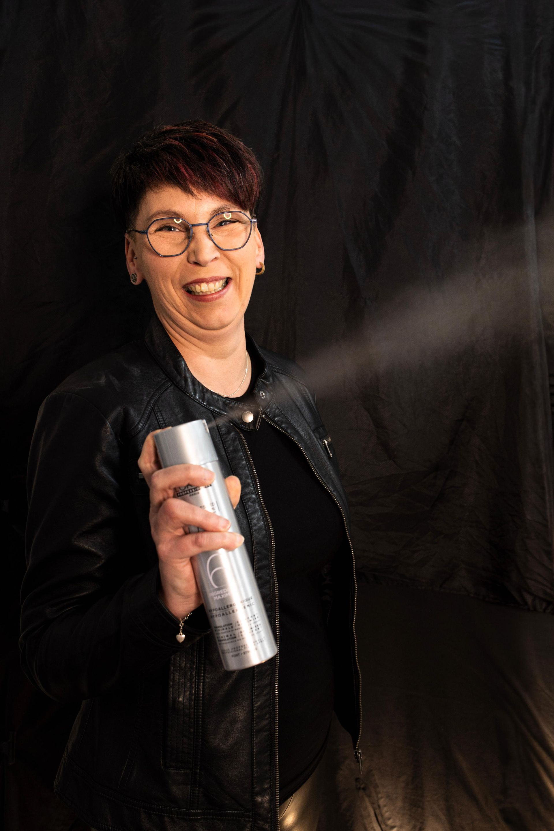 Frau lächelt, sprüht Haarspray. Dunkler Hintergrund, trägt Brille und Lederjacke, hält eine silberne Dose.