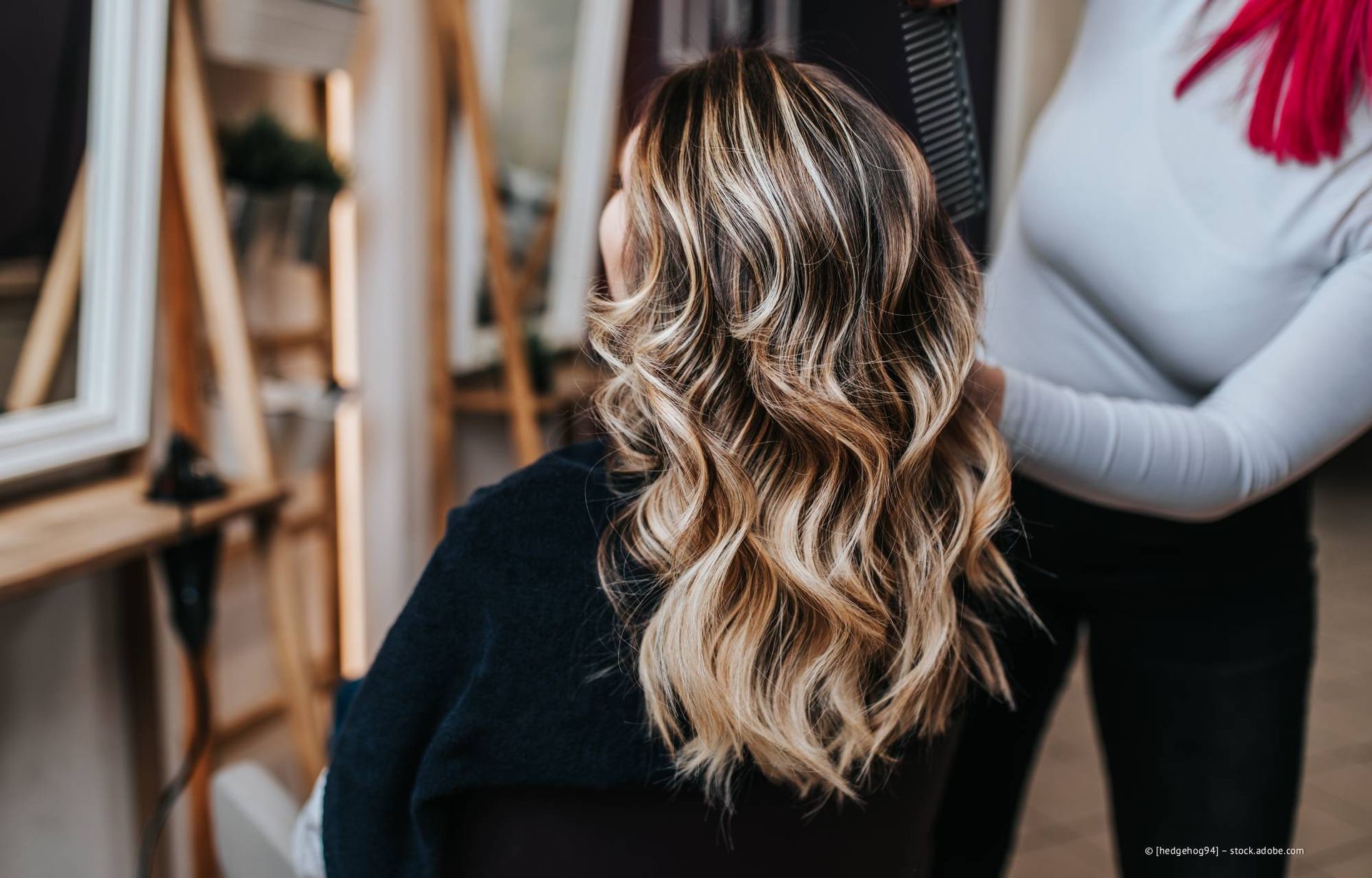 Frau mit welligem, gesträhntem Haar auf einem Friseurstuhl, Stylistin berührt ihr Haar.
