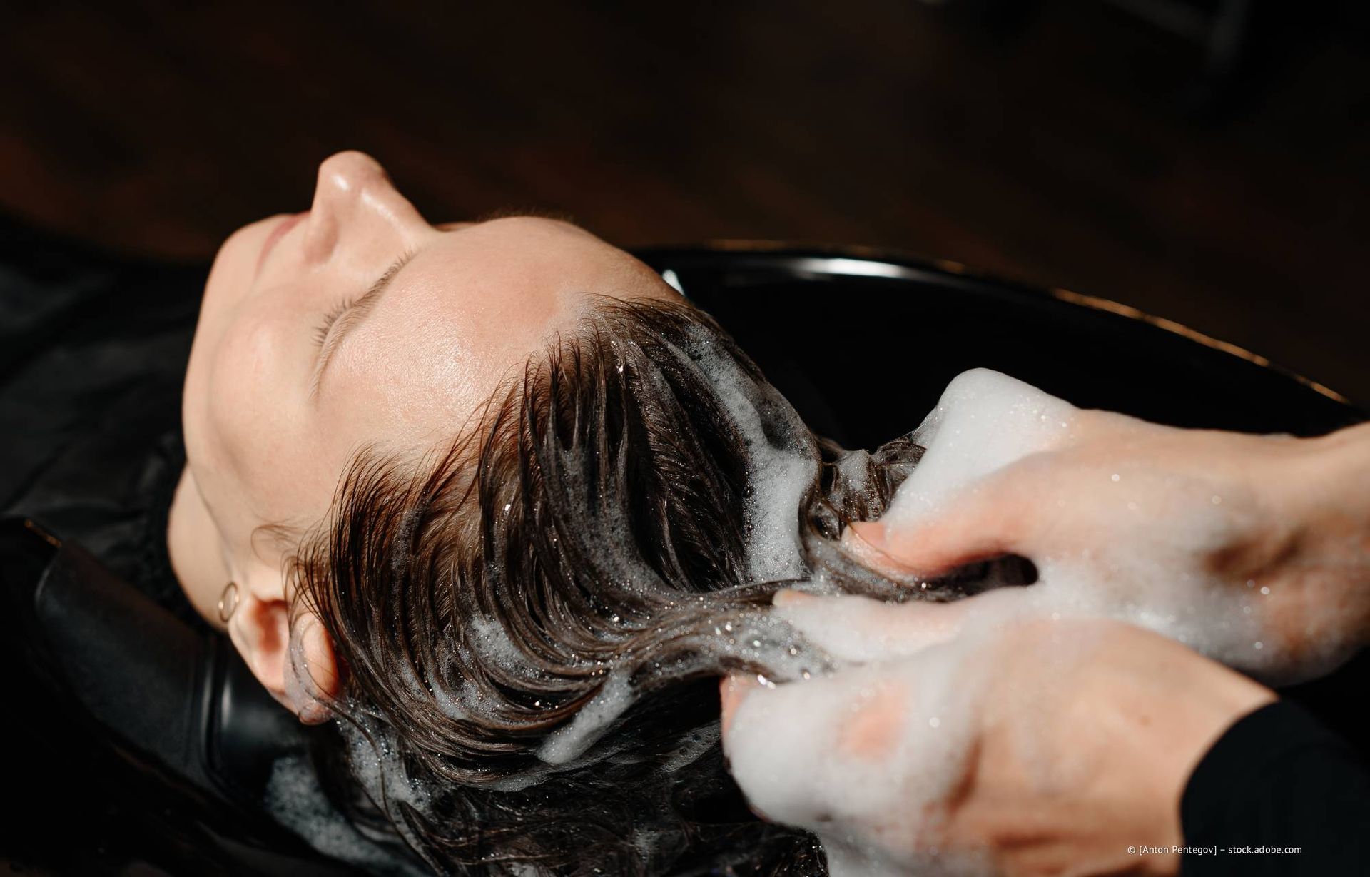 Eine Person lässt sich in einem Salonwaschbecken die Haare waschen; Seifenreste im Haar und an den Händen.