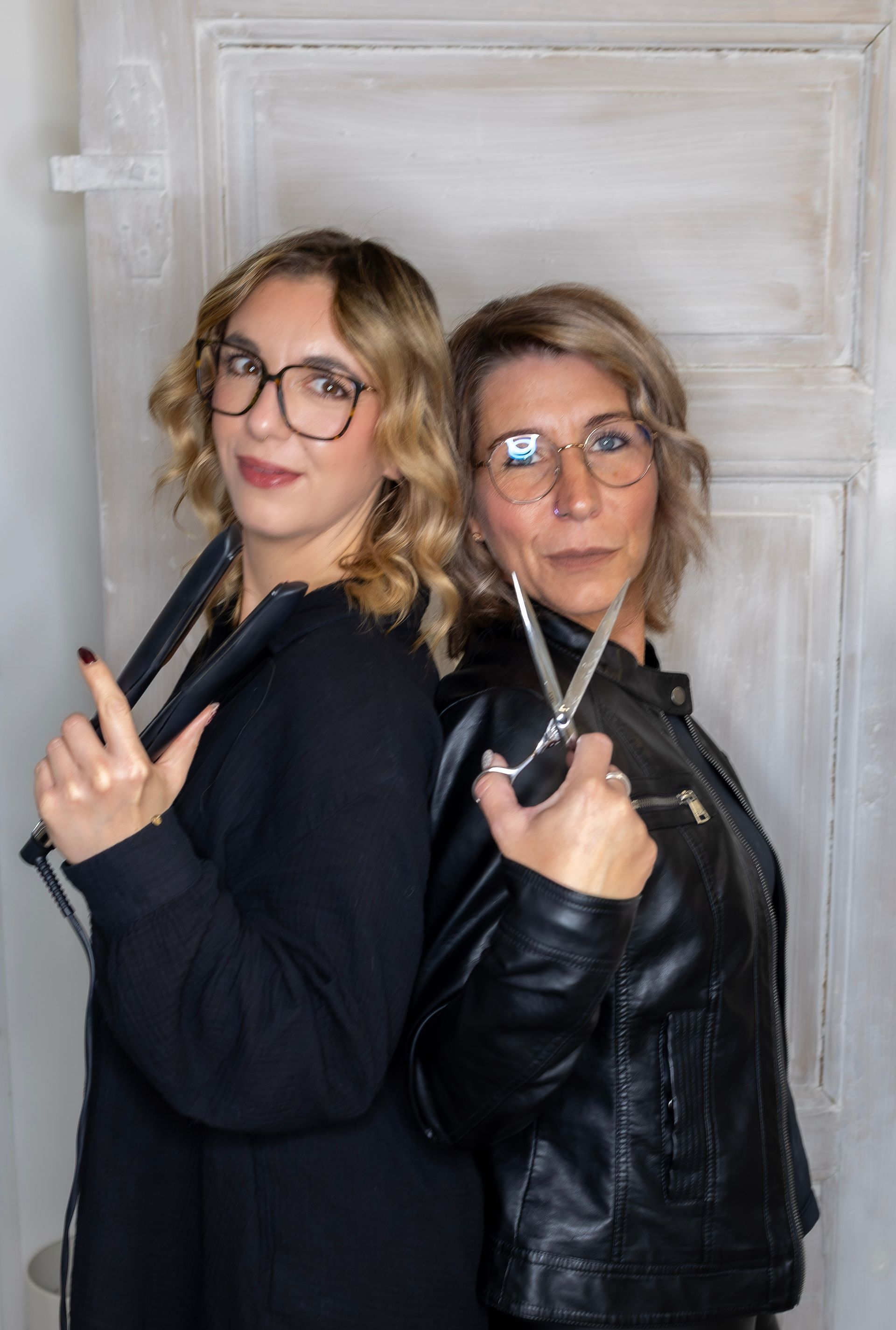 Zwei Frauen mit Brille und Schere stehen Rücken an Rücken. Die eine trägt ein schwarzes Outfit, die andere eine schwarze Lederjacke.