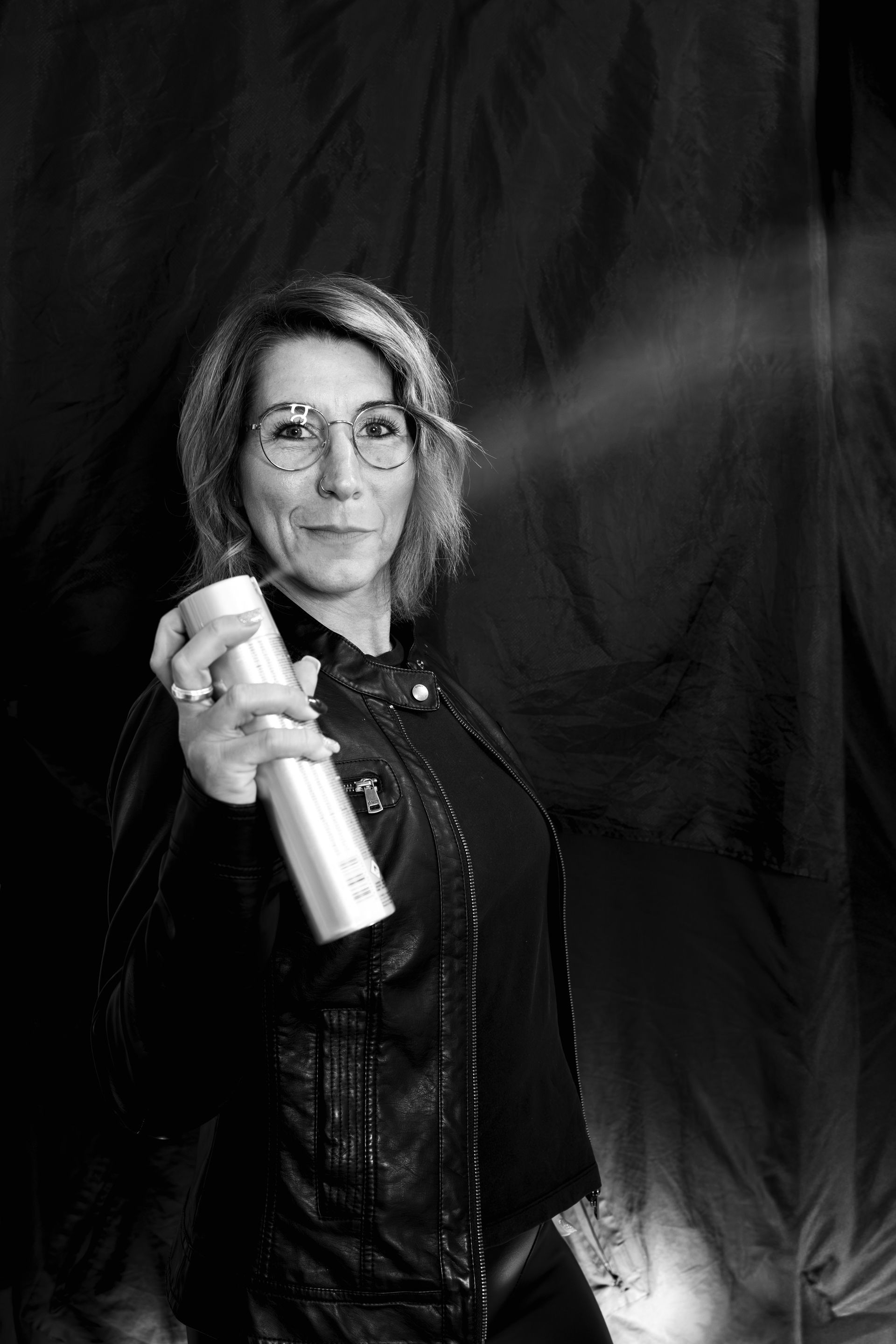 Frau hält Haarspray in der Hand und sprüht in Richtung Kamera; dunkler Hintergrund.