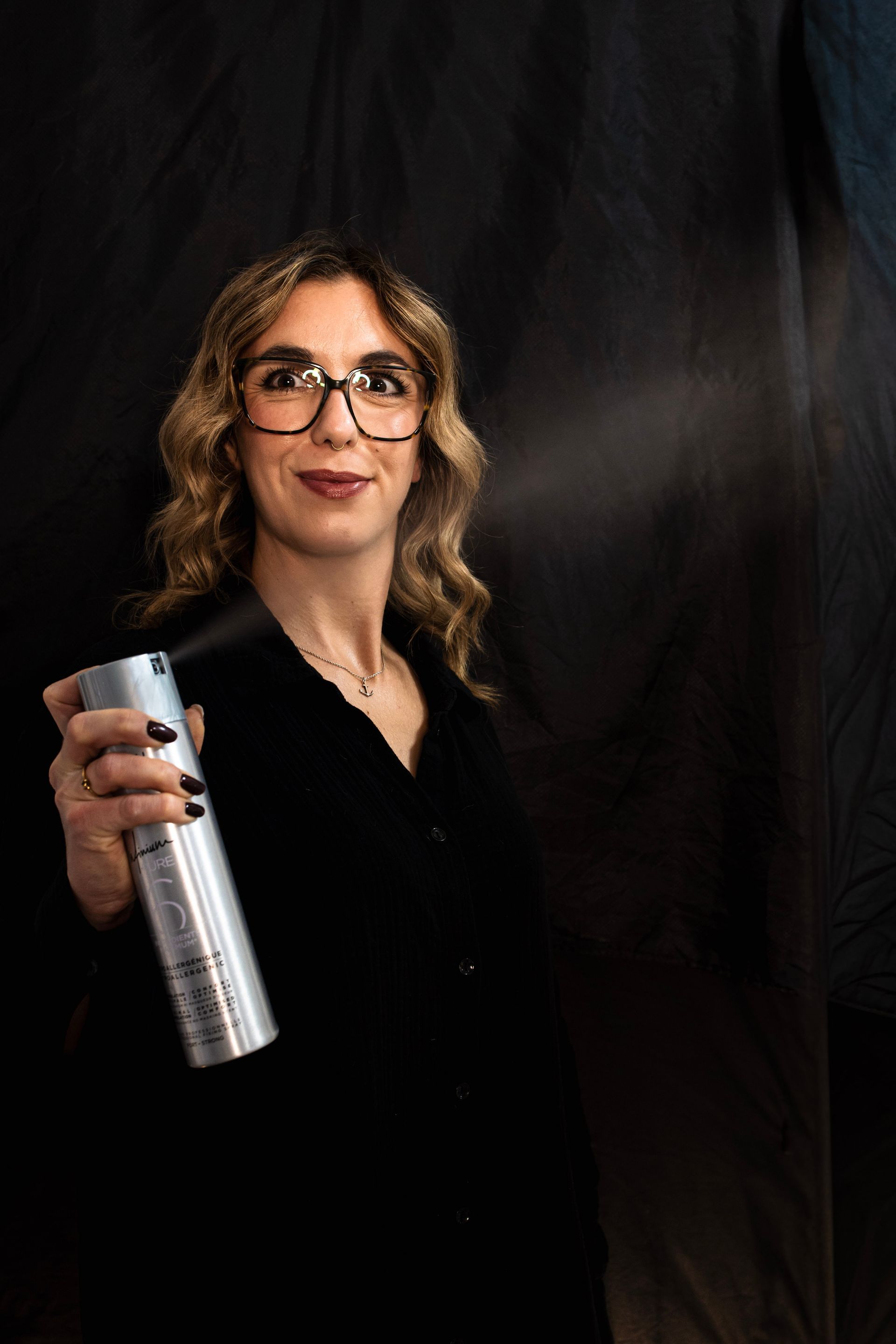 Frau sprüht Haarspray auf, hält eine silberne Dose in der Hand, dunkler Hintergrund.